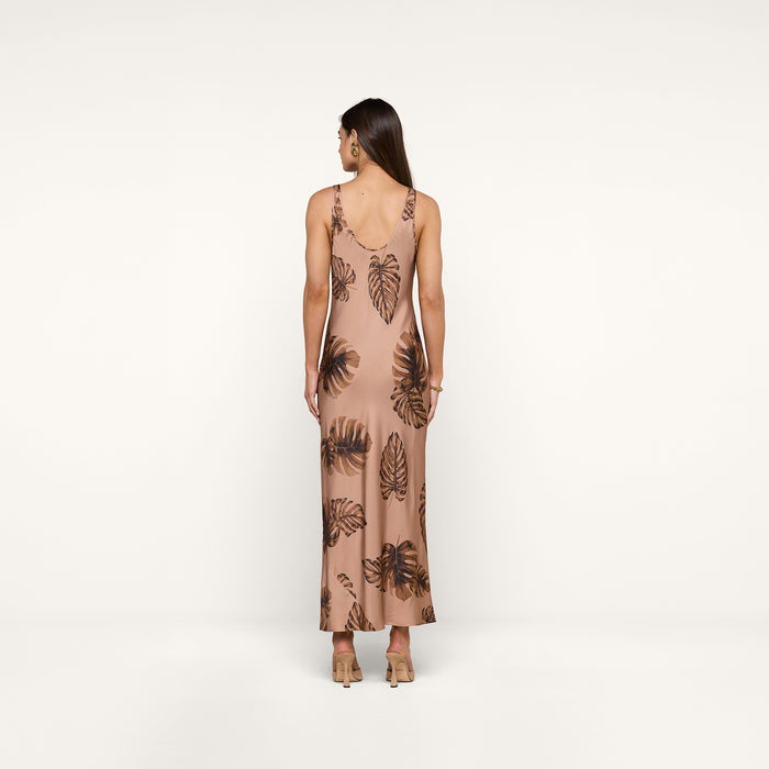 A woman stands with her back to the camera, wearing the Akiya Satin Maxi Dress—a long, sleeveless, bias-cut dress in a brown palm leaf print. She has straight brown hair and is accessorized with beige heels and hoop earrings. The background is plain white.