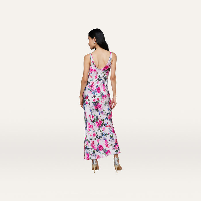A woman faces away, wearing the Akiya Satin Maxi Dress—a long, sleeveless, bias-cut dress with a pink rose print—and beige high heels, standing against a plain light background.