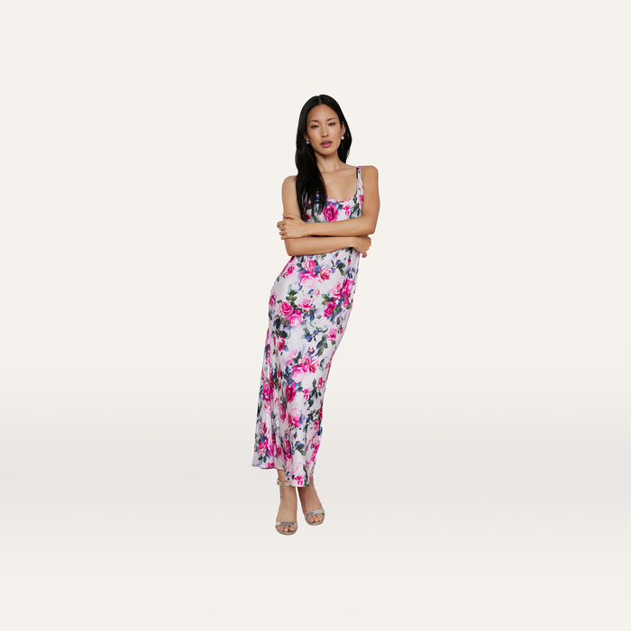 A woman stands with her arms crossed against a plain white background, wearing the Akiya Satin Maxi Dress—a long, sleeveless dress featuring a pink rose print adorned with purple and white flowers.