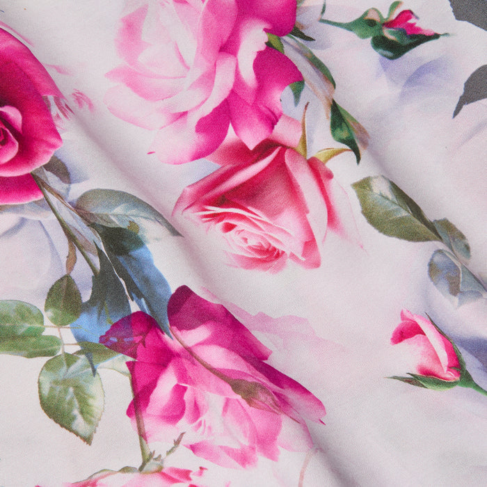 A close-up of the Akiya Satin Maxi Dress showcases its pink rose print with green leaves on a white background. The satin material drapes in gentle folds, adding texture and depth to the floral design.