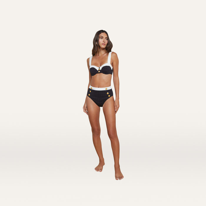A woman stands barefoot against a plain white background wearing the Alexandria Bikini Top, a black and white high-waisted bikini crafted from textured bouclé fabric with gold button details and featuring an underwire balconette design.