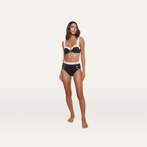 A woman stands barefoot against a plain white background wearing the Alexandria Bikini Top, a black and white high-waisted bikini crafted from textured bouclé fabric with gold button details and featuring an underwire balconette design.