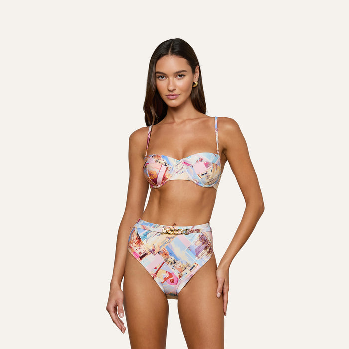 A woman stands against a plain white background, facing forward with a relaxed expression, wearing the Alexandria Bikini Top—a colorful, high-waisted bikini set featuring a postcard print and offering UPF 50 protection through its balconette-style design.
