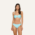A woman stands against a plain light background, wearing the Alexandria Bikini Top in ice blue with a neutral expression.