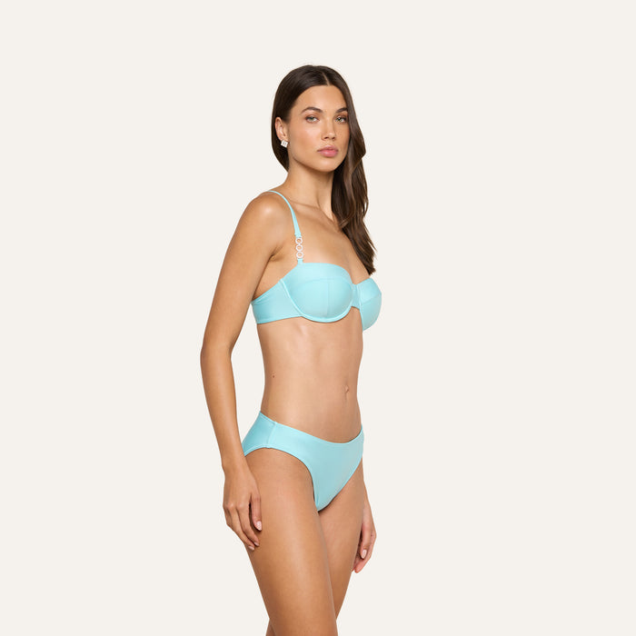 A woman stands against a plain background, wearing the Alexandria Bikini Top in ice blue with adjustable straps. The balconette design offers a flattering fit. She has long brown hair, a neutral expression, and her body is angled slightly to the side.
