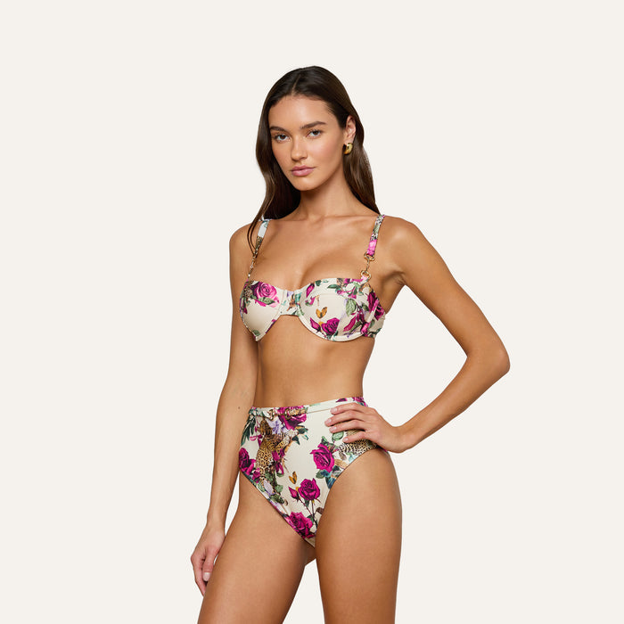 A woman poses in the Alexandria Bikini Top, a white balconette style with rose leopard print featuring underwire and adjustable straps. She pairs it with matching high-waisted bottoms, standing against a plain light background.