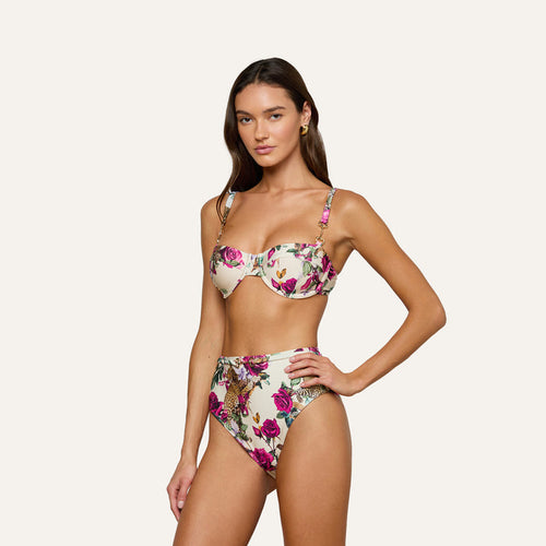 A woman poses in the Alexandria Bikini Top, a white balconette style with rose leopard print featuring underwire and adjustable straps. She pairs it with matching high-waisted bottoms, standing against a plain light background.