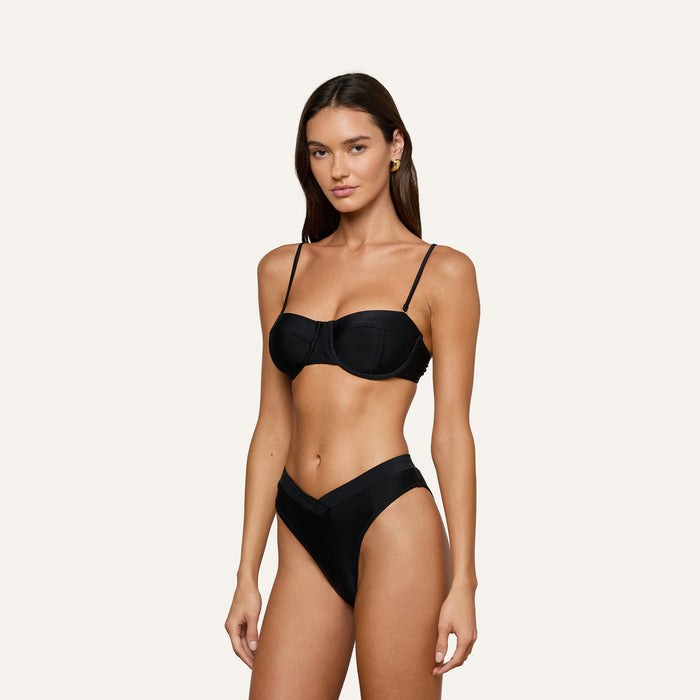 A woman wearing the Alexandria Bikini Top, a black underwire balconette bikini, paired with V-cut high-waisted bottoms, stands against a plain light background.
