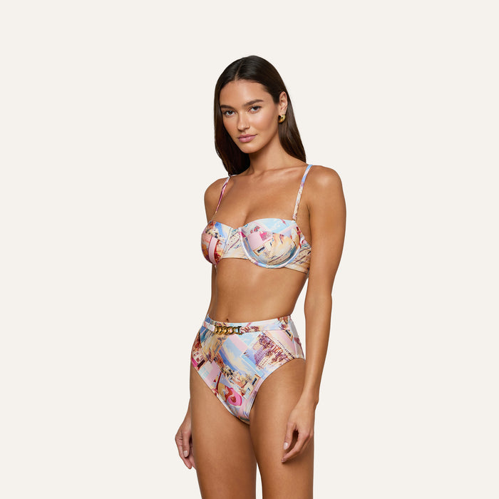 A woman wearing the Alexandria Bikini Top with high-waisted, colorful postcard print bottoms stands against a plain, light background, looking at the camera with a relaxed expression.