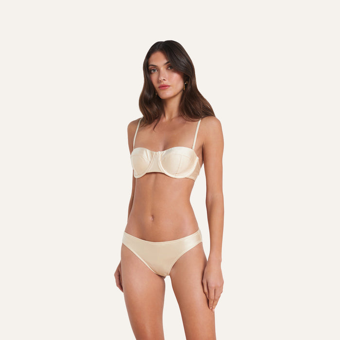A woman stands against a plain background, wearing the Alexandria Bikini Top—a cream-colored underwire bikini with adjustable straps—paired with matching panties.