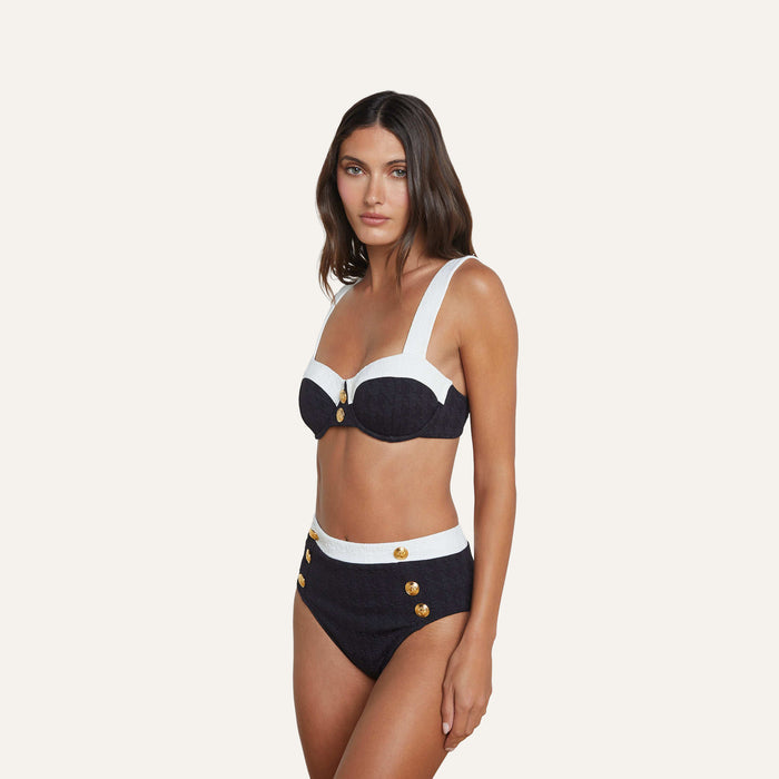 A woman poses against a plain light background, wearing the Alexandria Bikini Top—a navy and white balconette design with gold button details, underwire support, thick shoulder straps, and crafted from textured bouclé fabric. Paired with high-waisted bottoms.