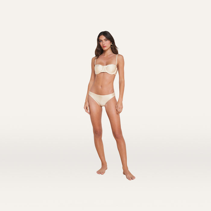 A woman stands barefoot against a plain white background, wearing the Alexandria Bikini Top with matching cream-colored bottoms, both featuring adjustable straps.
