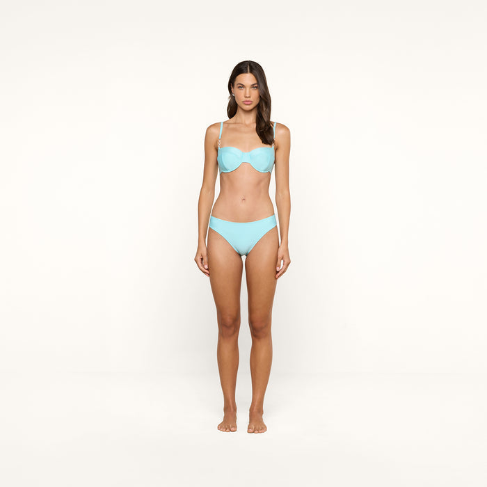 A woman stands barefoot facing forward, wearing an ice blue Alexandria Bikini Top as part of her bikini set, against a plain white background.
