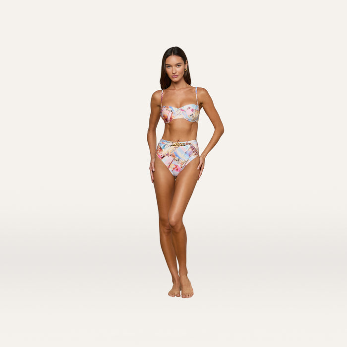 A woman with long, straight hair and a relaxed expression stands barefoot against a plain background, wearing the Alexandria Bikini Top with high-waisted bottoms. The colorful postcard print swimsuit features a balconette bikini top and offers UPF 50 protection.