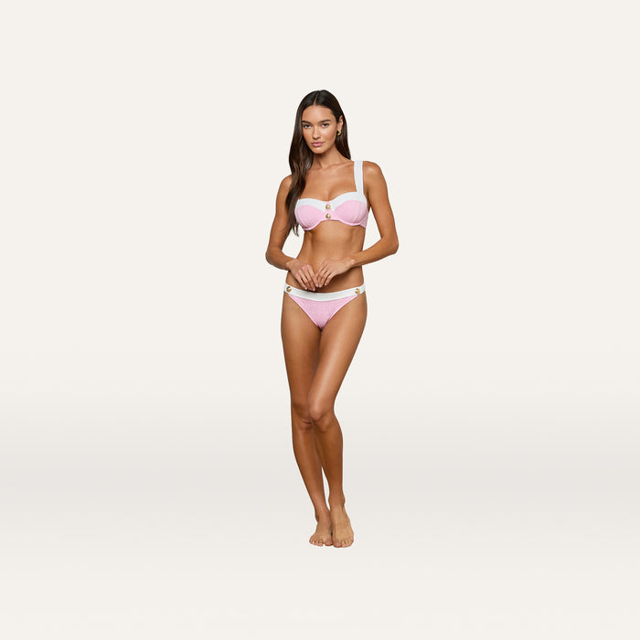 A woman stands barefoot on a plain white background, wearing the Alexandria Bikini Top in light pink with gold accents and textured bouclé fabric. She faces forward with her arms relaxed by her sides.