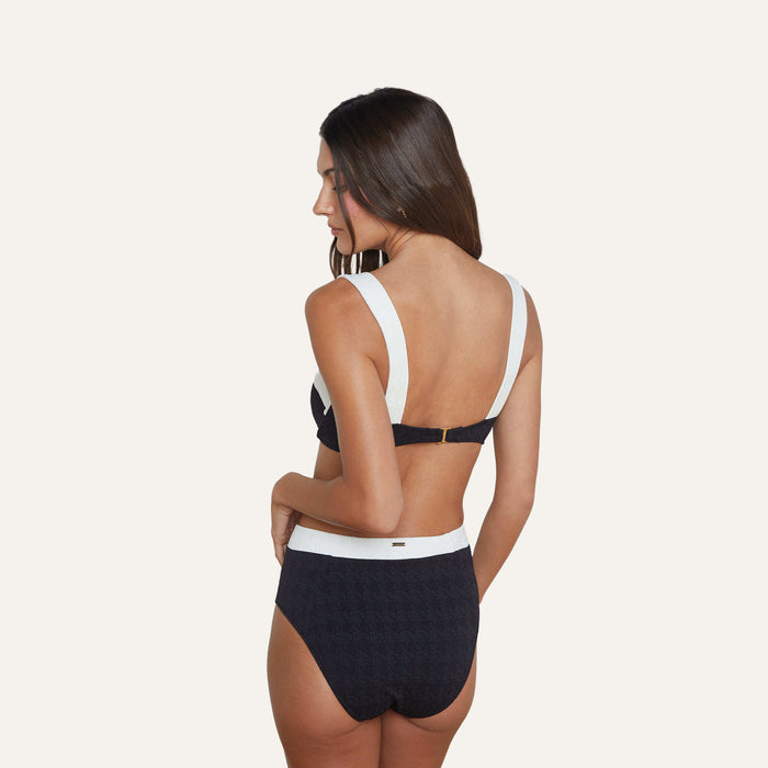 A woman with long brown hair stands with her back to the camera, wearing the Alexandria Bikini Top—a black and white balconette style with underwire—and coordinating high-waisted bottoms, both crafted from textured bouclé fabric, set against a plain light background.