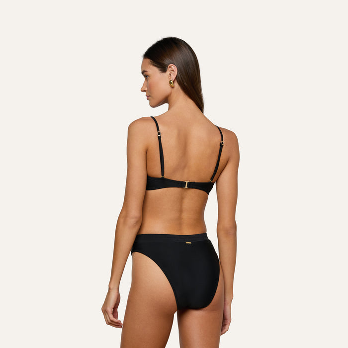 A woman stands with her back to the camera against a plain light background, wearing the Alexandria Bikini Top—a black balconette style featuring gold accents.