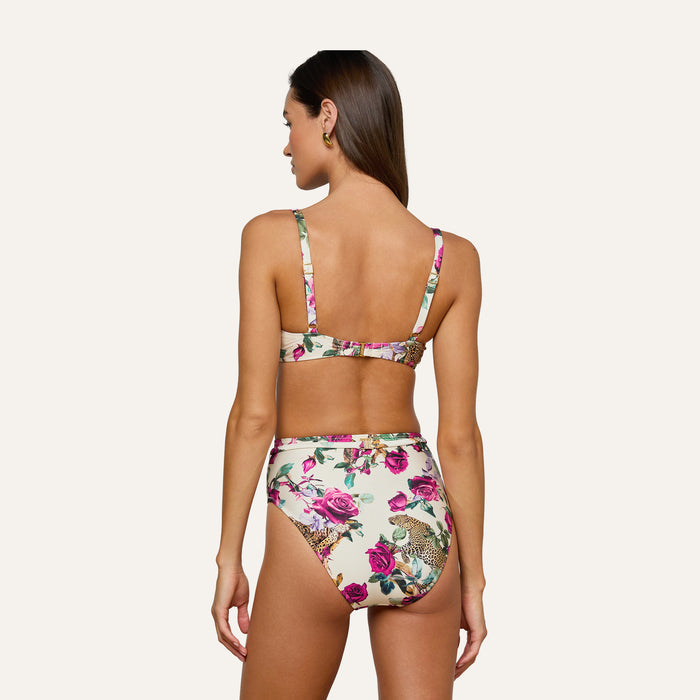 A woman with long brown hair is seen from the back wearing the Alexandria Bikini Top, which features pink and green floral patterns. She stands against a plain, light background.