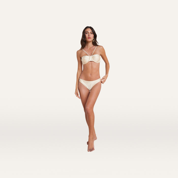 A woman stands facing forward, wearing the Alexandria Bikini Top in cream with adjustable straps and crossed details, against a plain white background.