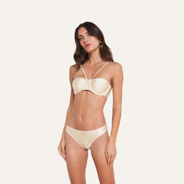 A woman wearing the Alexandria Bikini Top—a cream-colored, silk-infused style with thin adjustable straps—paired with hoop earrings, stands against a plain white background.