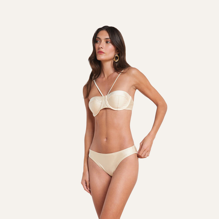 Woman standing against a plain background, wearing the Alexandria Bikini Top—a cream-colored, underwire bikini top with minimal straps—paired with gold hoop earrings.