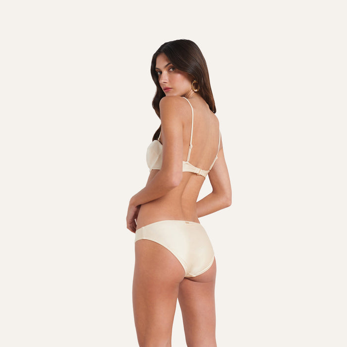 A woman with long brown hair stands turned to the side, wearing the Alexandria Bikini Top—a matching light beige underwire bikini top with adjustable straps—paired with a coordinating underwear set, against a plain white background.