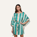 A woman wearing the Alia Cover-Up Tunic in a short, turquoise and white pattern with gold accents, wide sleeves, and a fabric belt at the waist stands against a plain background.