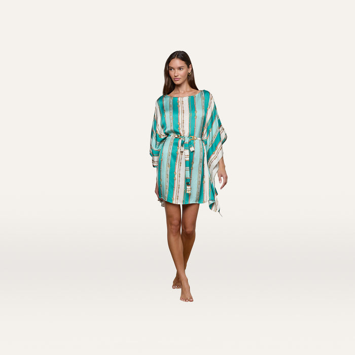 A woman wearing the Alia Cover-Up Tunic—a turquoise, white, and gold belt-stripe print dress with a matching belt—stands barefoot against a plain white background.