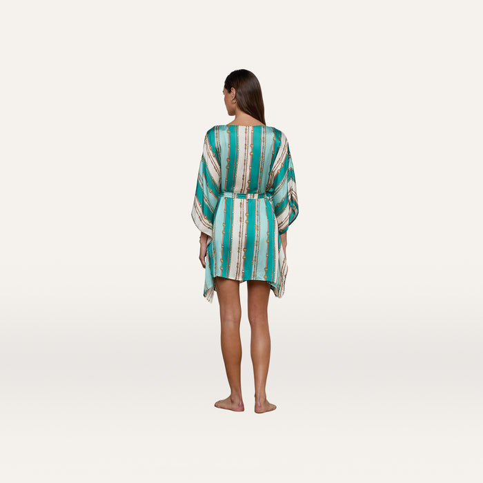 A woman stands barefoot facing away against a plain white background, wearing the Alia Cover-Up Tunic—a short, loose design with long sleeves and a turquoise and white belt-stripe print.
