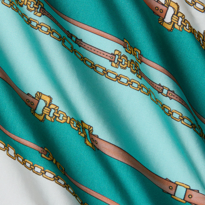 The Alia Cover-Up Tunic features silky fabric with a belt-stripe print of brown belts and gold chains on a teal and white background, displayed with soft folds and luminous highlights.