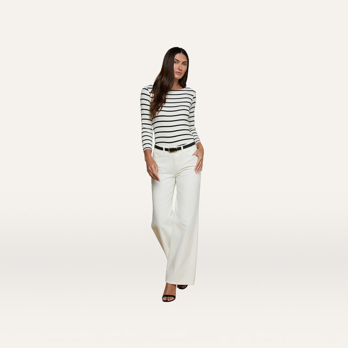 A woman stands against a plain background, wearing a black and white striped long-sleeve top, high rise Alicent Wide-Leg Jeans in white, a black belt, and black heels.
