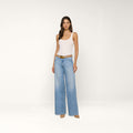 A woman stands against a plain white background, wearing a white tank top, the Alicent Wide-Leg Jean in light blue with a brown belt, and beige heels.