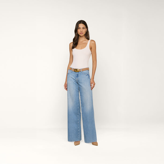 A woman stands against a plain white background, wearing a white tank top, the Alicent Wide-Leg Jean in light blue with a brown belt, and beige heels.