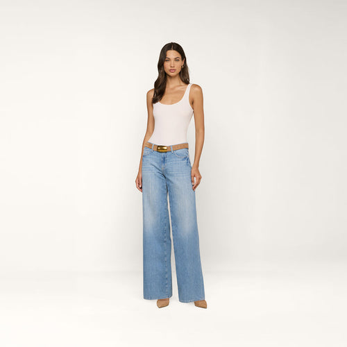 A woman stands against a plain white background, wearing a white tank top, the Alicent Wide-Leg Jean in light blue with a brown belt, and beige heels.