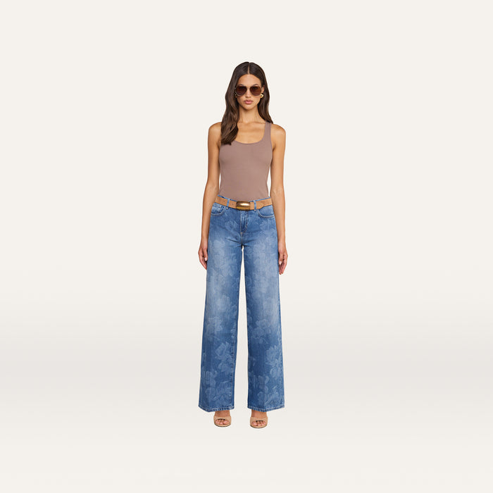 Woman standing against a plain background, wearing sunglasses, a taupe tank top, the Alicent Wide-Leg Jean—an all-cotton, sneaker-length denim style with a subtle floral print—paired with a tan belt and open-toe heels.