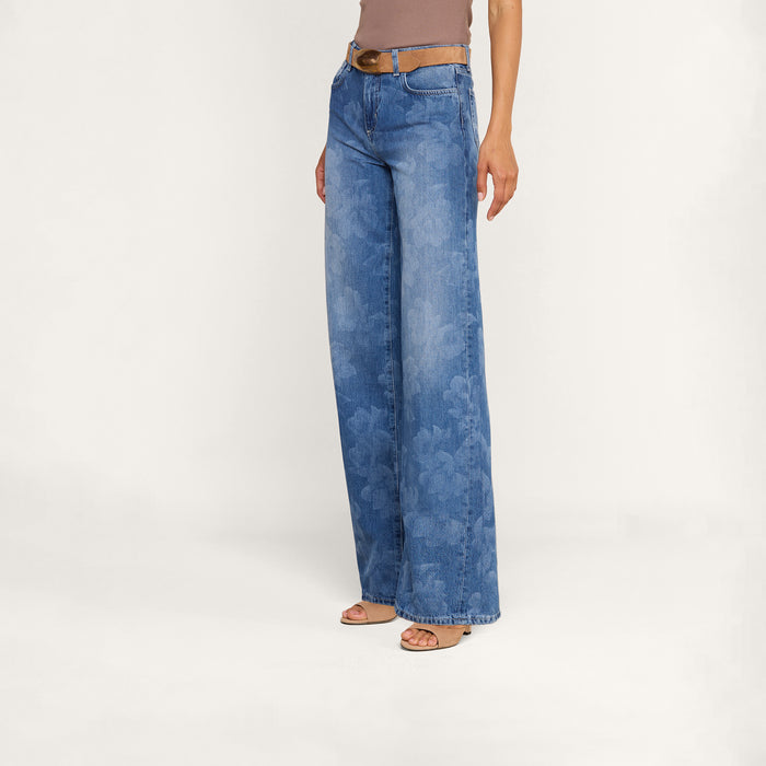 Wearing the Alicent Wide-Leg Jean—crafted from all-cotton denim with a subtle floral print—paired with a brown belt, taupe sleeveless top, and sandals, this person stands against a plain white background.