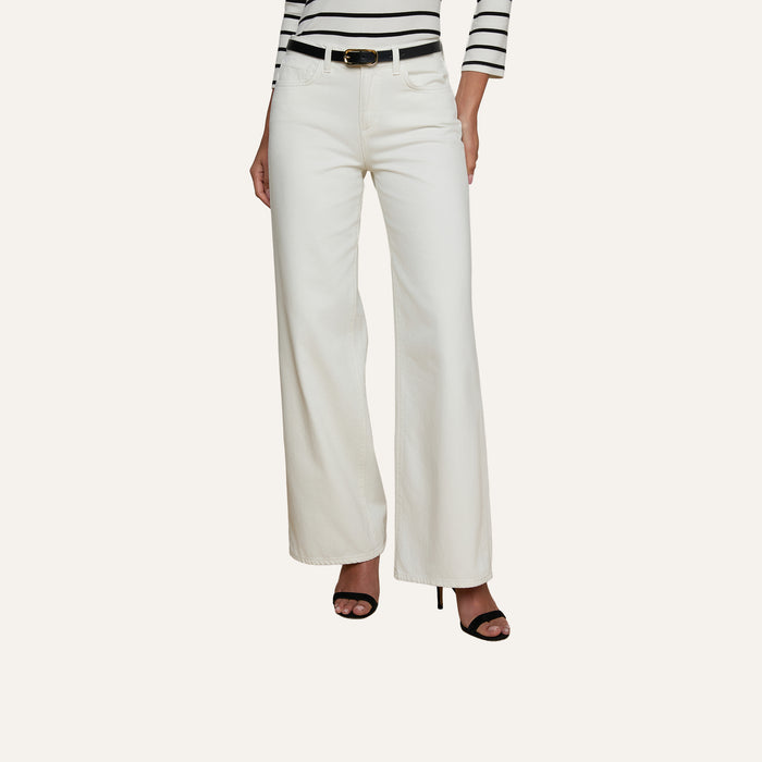 A woman is shown from the shoulders down wearing the Alicent Wide-Leg Jean in white with a high-rise fit, styled with a black belt, black high-heeled sandals, and a black-and-white striped long-sleeve top. The background is plain and light-colored.
