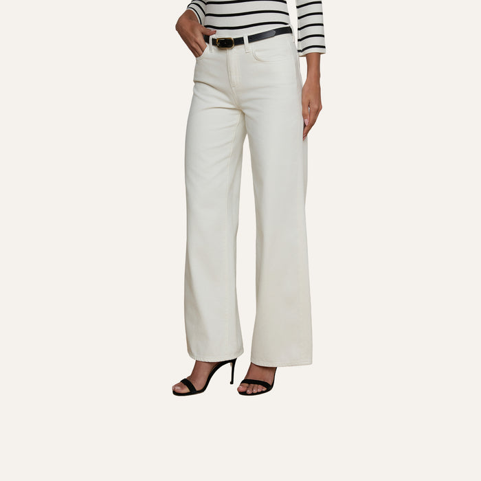 A woman stands against a plain light background wearing the Alicent Wide-Leg Jean in white with a high rise, paired with a black belt, a black and white striped long-sleeve top, and black high-heeled sandals. Only her body is visible; her face is not shown.