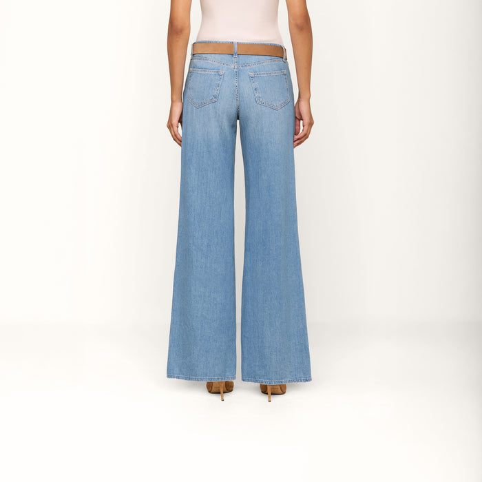 A person stands facing away against a plain white background, wearing the Alicent Wide-Leg Jean in light blue, styled with a tan belt, a light tank top, and tan heeled shoes.
