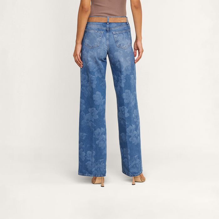 A person wearing the Alicent Wide-Leg Jean in a sneaker-length, high-waisted blue floral print, paired with a taupe sleeveless top and beige heeled sandals, shown from the back against a plain light background.