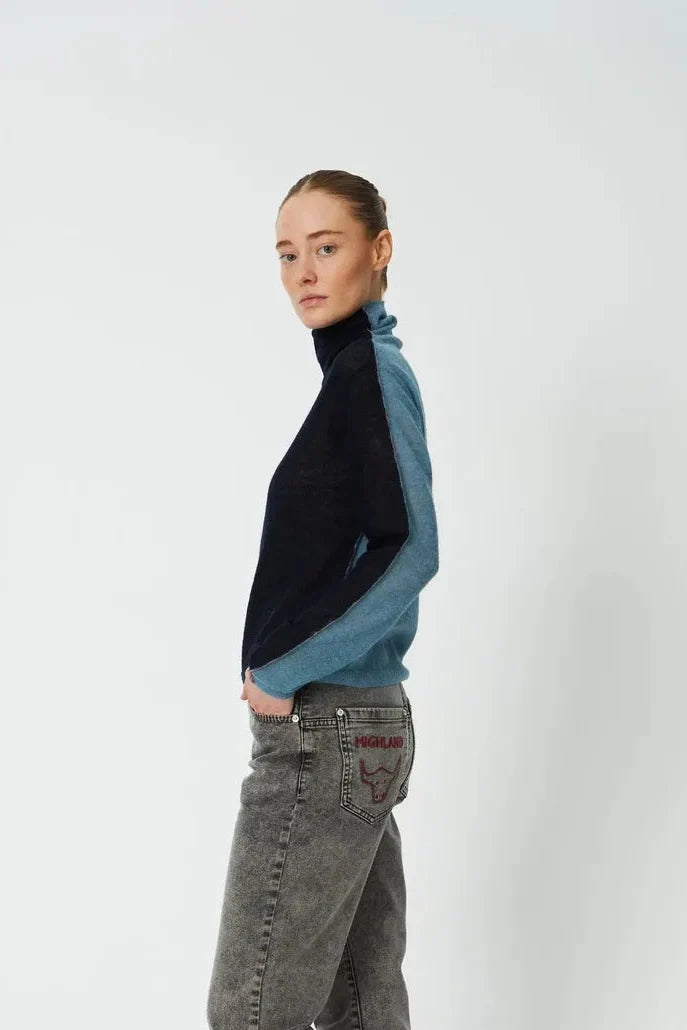 A model stands in profile wearing a color-blocked knit turtleneck sweater with black and blue panels, gray jeans, and red slip-on shoes against a plain light gray studio background.