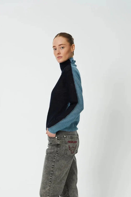 A model stands in profile wearing a color-blocked knit turtleneck sweater with black and blue panels, gray jeans, and red slip-on shoes against a plain light gray studio background.