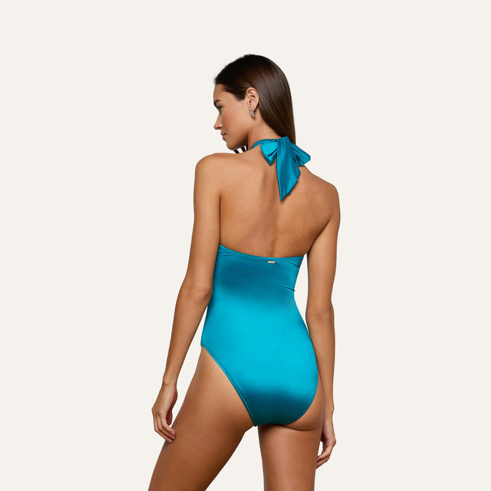 A woman with her hair down stands sideways against a plain light background, looking over her left shoulder while wearing the Althea Bandeau One-Piece Swimsuit in bright blue—an elegant, backless halter style that offers UPF 50 swimwear protection.