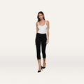 Wearing the Amaris Pedal Pusher in black with a white tank top and black heels, a woman stands against a plain light background, walking forward with a neutral expression.