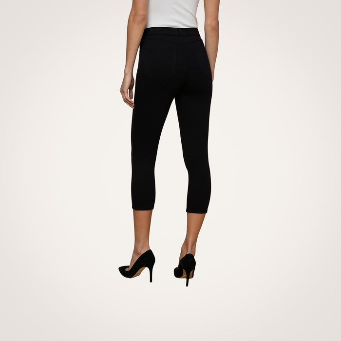 A person is seen from the back, standing against a plain light background, wearing a white sleeveless top and the Amaris Pedal Pusher—black ponte knit pants with a body-hugging fit—paired with black high heels.