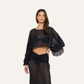 A woman poses against a plain light background wearing the Ambala Mesh Cover-Up Top, a sheer black long-sleeve crop top that creates a chic, cropped silhouette. She pairs it with a matching sheer black skirt, accessorizes minimally, and lets her long brown hair fall naturally as she maintains a neutral expression.
