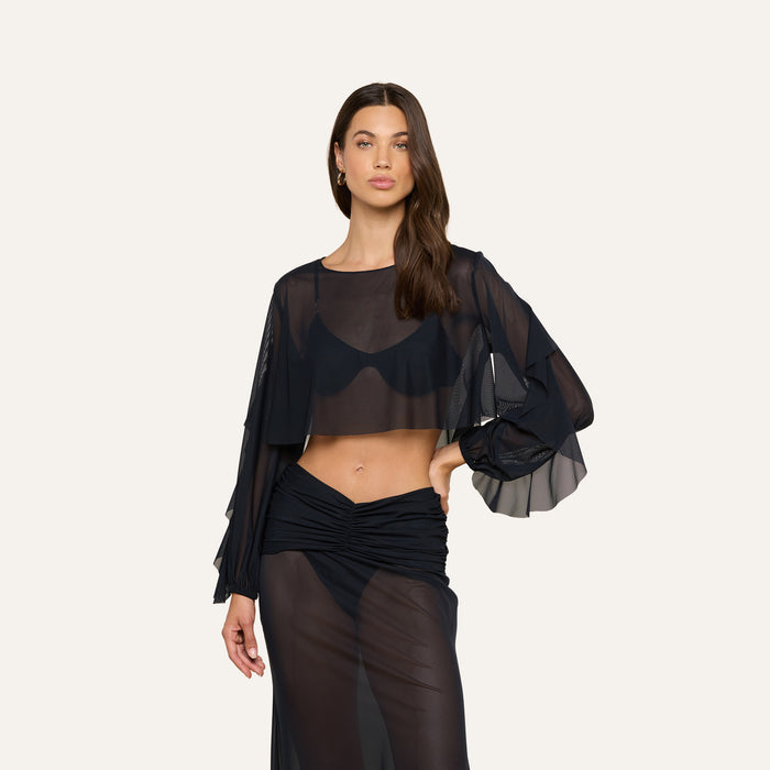 A woman poses against a plain light background wearing the Ambala Mesh Cover-Up Top, a sheer black long-sleeve crop top that creates a chic, cropped silhouette. She pairs it with a matching sheer black skirt, accessorizes minimally, and lets her long brown hair fall naturally as she maintains a neutral expression.