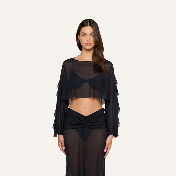 A woman wearing the Ambala Mesh Cover-Up Top— a sheer black long-sleeve crop top with ruffled sleeves—paired with a matching sheer black skirt, shows off a relaxed beachside-inspired look against a plain light background.