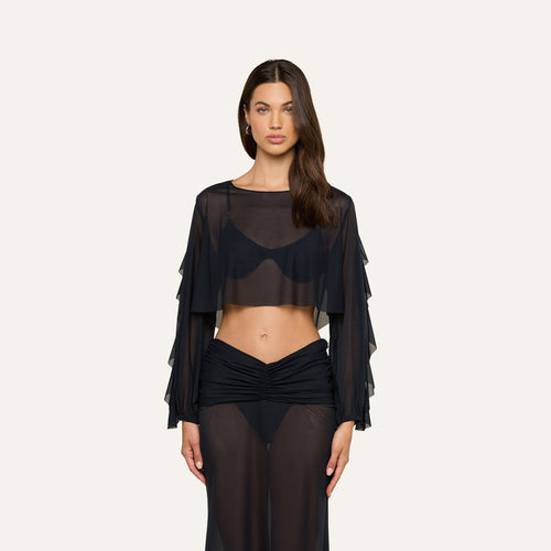 A woman wearing the Ambala Mesh Cover-Up Top— a sheer black long-sleeve crop top with ruffled sleeves—paired with a matching sheer black skirt, shows off a relaxed beachside-inspired look against a plain light background.