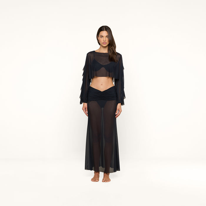 A woman stands barefoot against a plain white background, wearing the Ambala Mesh Cover-Up Top—a sheer black long-sleeve piece with a cropped silhouette—paired with a matching sheer black skirt over black undergarments, evoking relaxed beachside style.